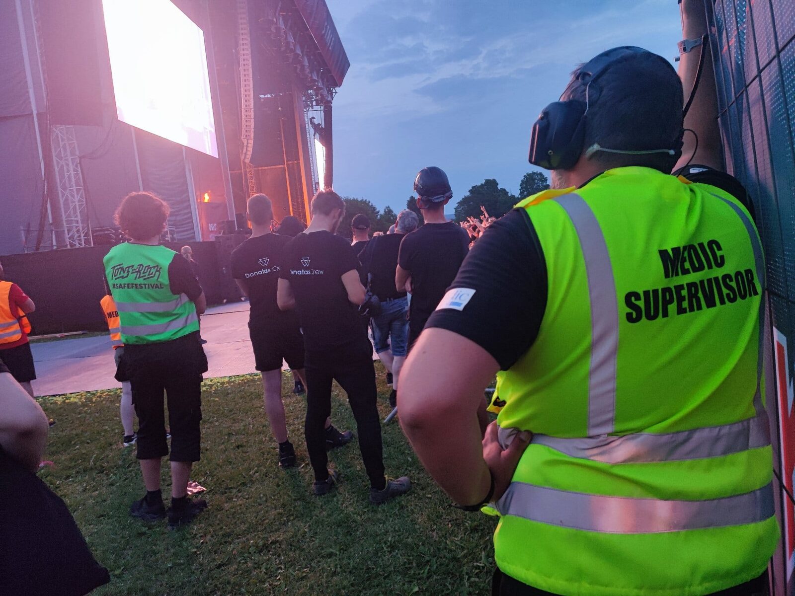A Medic Supervisor in a yellow vest and headset stands with his back to the camera in front of the stage at Tons of Rock, looking out over the audience. Nothing has happened.