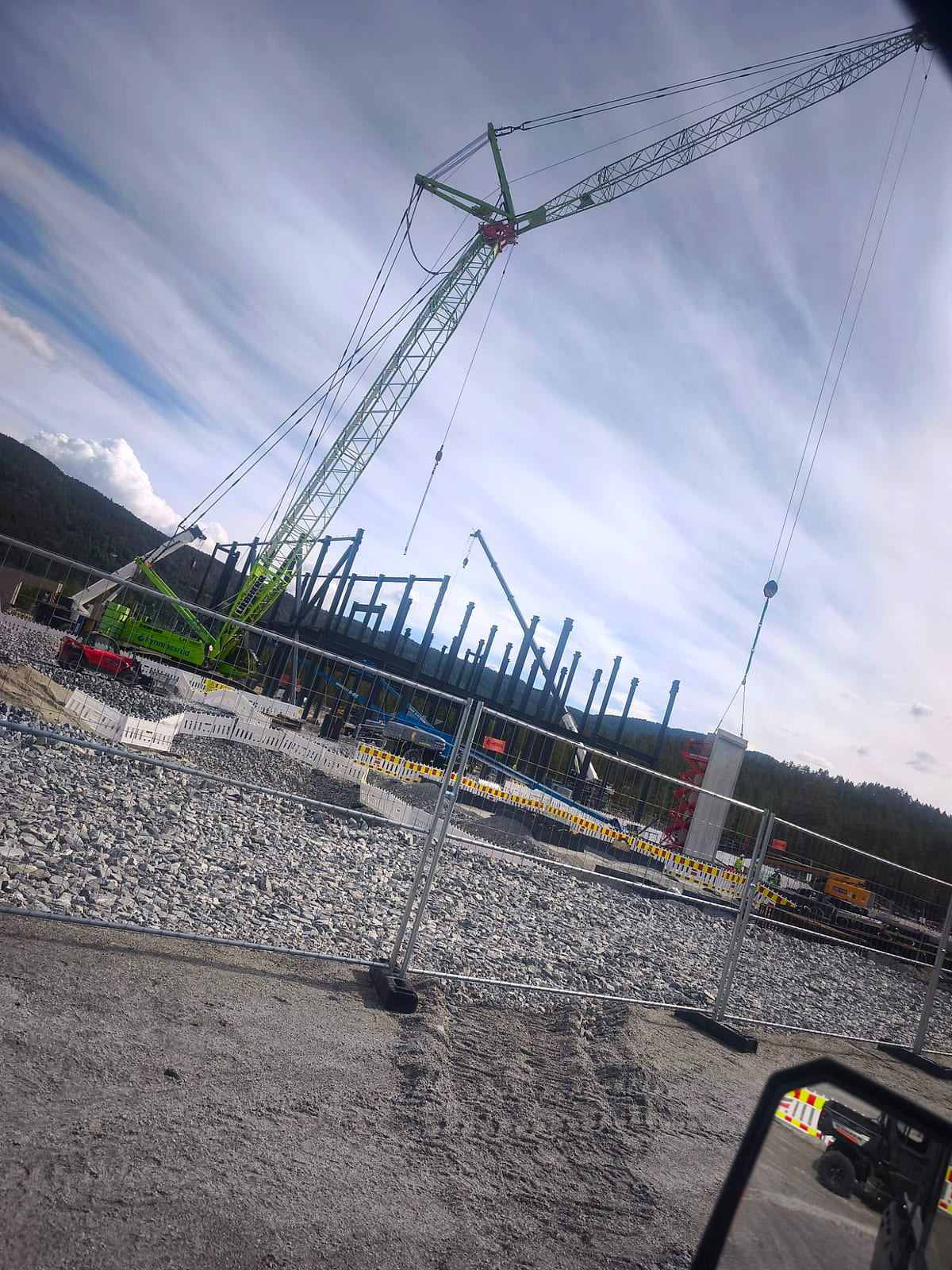 Construction site with a mobile crane and a steel frame being assembled. Blasted rock in the foreground and wooded hills behind.