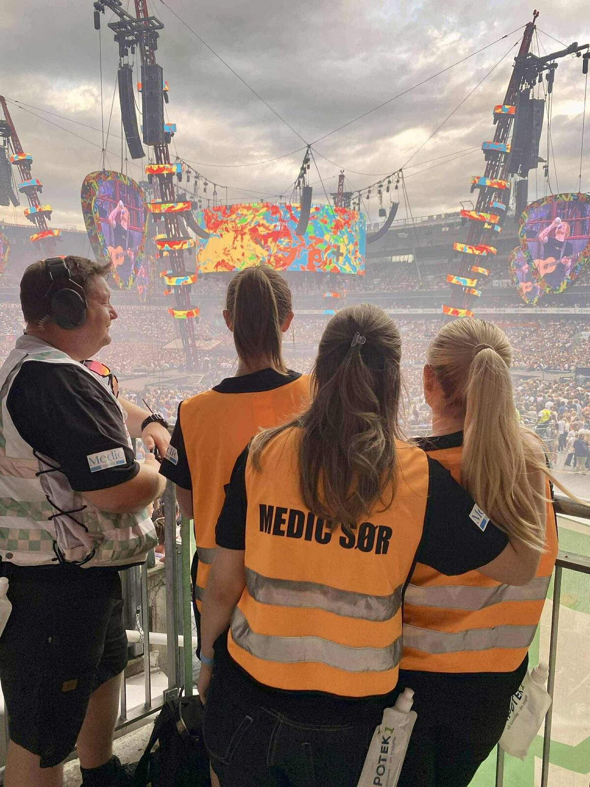 Four people from Medic Sør in orange vests stand with their backs to the camera on a stand, looking out over the audience at a stadium concert.