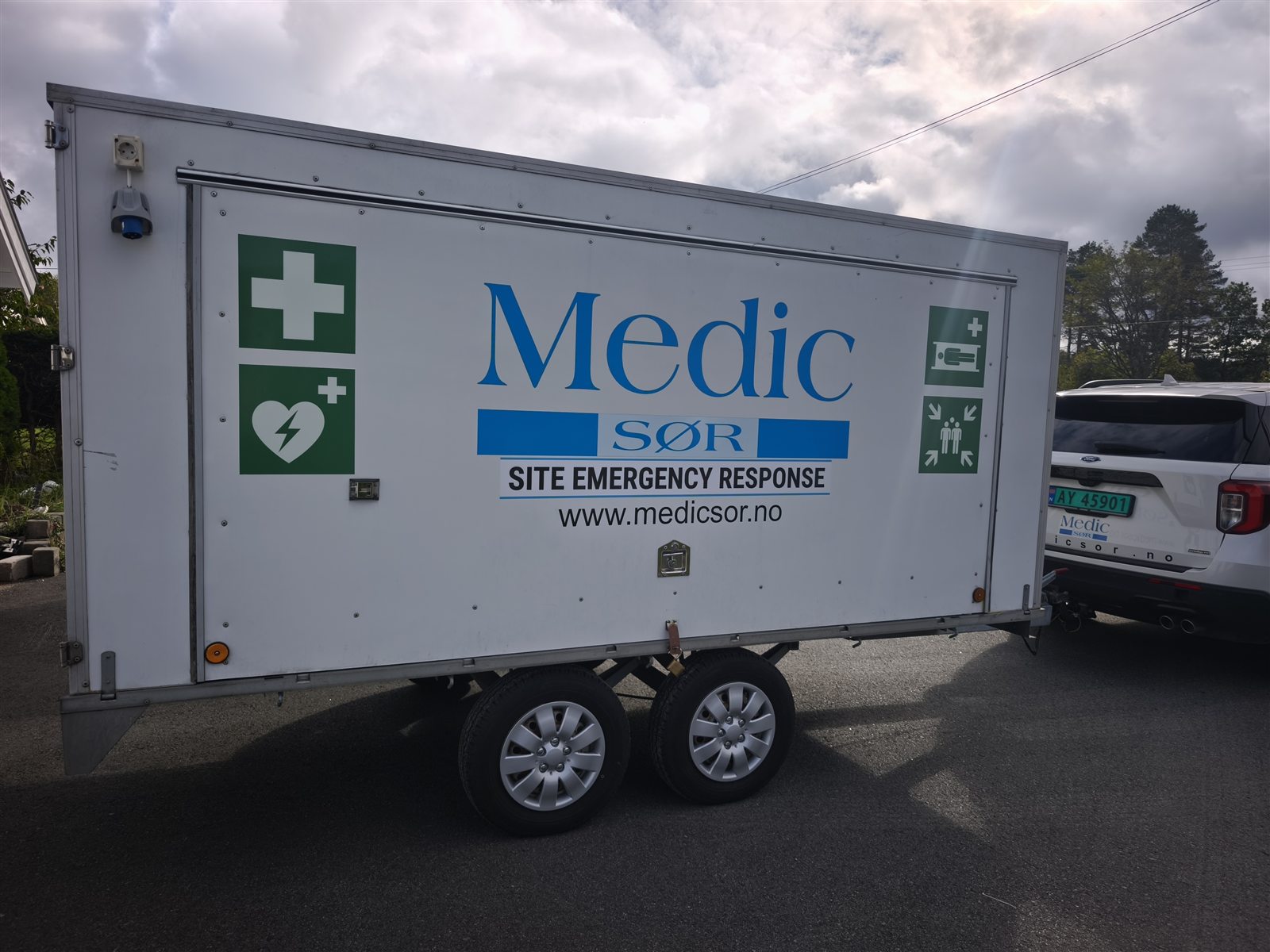 White Medic Sør trailer marked Site Emergency Response, with first aid and AED symbols, parked in front of a white car marked Medic Sør.